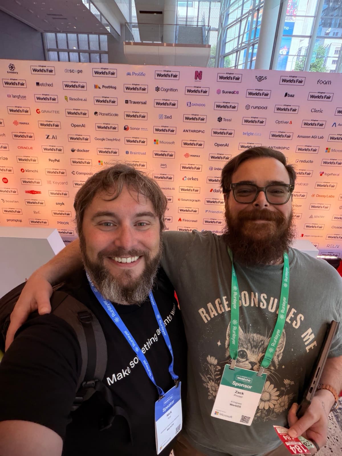 Nick Nisi and Zack Proser grinning in a selfie in front of the AI Engineer World's Fair sponsor wall
