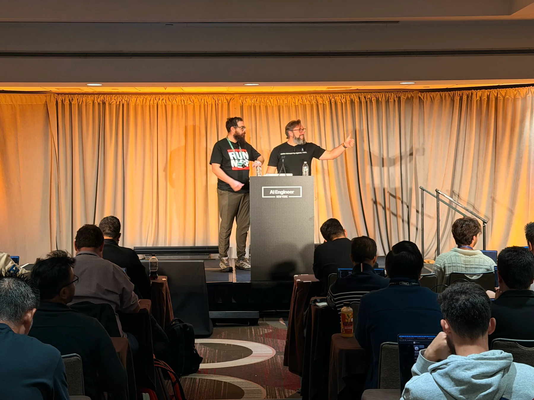 Zack speaking at the lectern with Nick in front of a seated audience at AI Engineer New York.